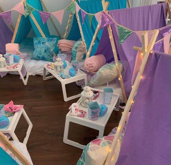 Indoor mermaid-themed sleepover setup with purple tents, adorned with string lights, small beds with cushions, and individual trays with sleep masks and lanterns.