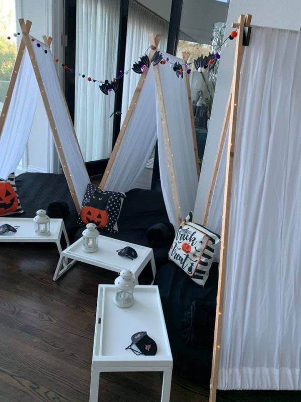 Indoor scene with two Lakeland teepee tent-like structures decorated with halloween-themed pillows, fairy lights, and garlands. there are white tables with lanterns and a halloween mask in front.