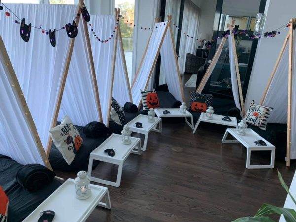 Indoor camping setup with small 246053091_1260052167752305_4680066096506698376_n tents adorned with halloween decorations, including bat garlands and pumpkin-themed pillows, arranged on a wooden floor. each tent has a small white side table.