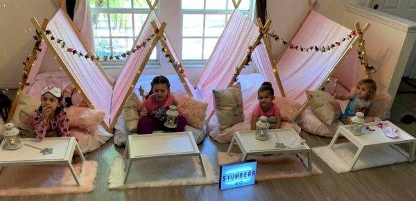 Four young girls at a slumber party, sitting inside individual Bell Tent teepees adorned with fairy lights, enjoying snacks on small trays in a cozy room.