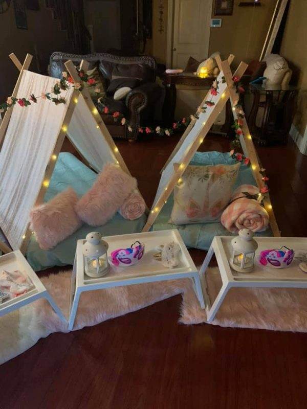 Three Lakeland teepees with pink and white decorations adorned with lights and flowers in a cozy room setting, accompanied by small tables with lanterns and plush pillows on a fur rug for the party.