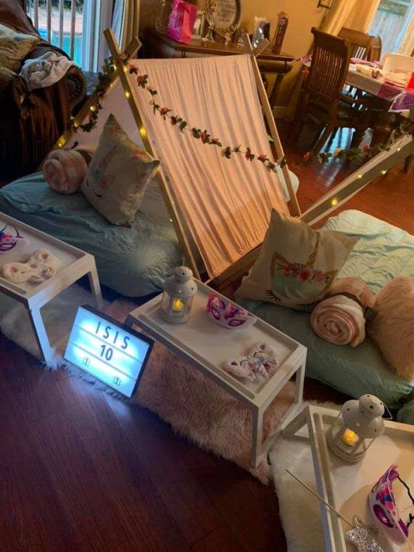 Cozy indoor tent made with a draped Unicorn Theme sheet over a frame, decorated with green garland and lights. floor cushions, small lanterns, a digital clock displaying "isis 10", and a tray of snacks enhance the comfortable setting.