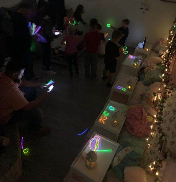 A dimly lit indoor room where children and adults play with glow stick bundles. Some participants are using glow stick bundles to make shapes while others watch, creating a festive atmosphere.