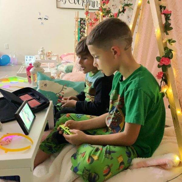 Two children sitting on a bed in a cozy, decorated room, focused on playing a video game on a handheld console with llama Theme. The room is softly lit with string lights and plush toys are visible.