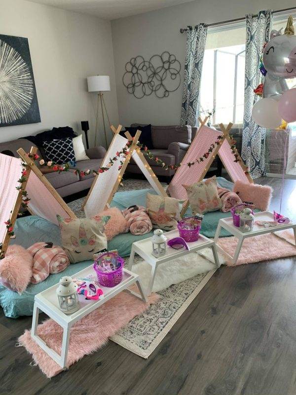 Cozy indoor sleepover setup featuring small wooden teepees with fairy lights, pastel cushions, blankets, lanterns, and a plush carpet in a living room decorated with the Unicorn Theme.