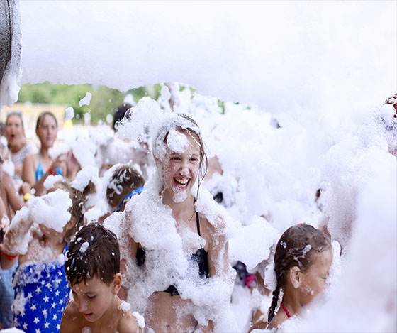 A joyful girl covered in Foam-Machine-extra-goody suds at a foam party, laughing amidst a crowd of people also enjoying the bubbly fun.