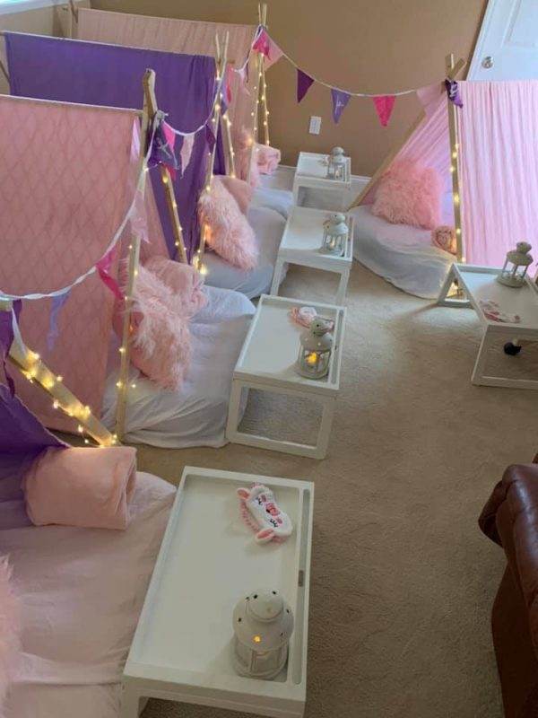 A cozy indoor sleepover setup with small white beds adorned with pink and white pillows, fluffy pink blankets, string fairy lights, and decorative dance Theme banners. each bed is paired with a small table hosting a lamp and slippers.