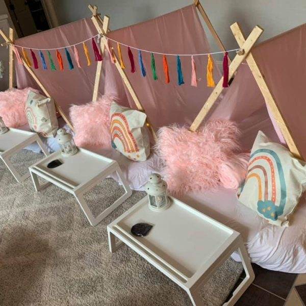 Indoor camping setup with three small tents made from Boho theme fabric, each featuring fluffy pink pillows, rainbow and cloud print cushions, accompanied by white trays in front of each tent. decorative string lights and multicolored flags hang above.