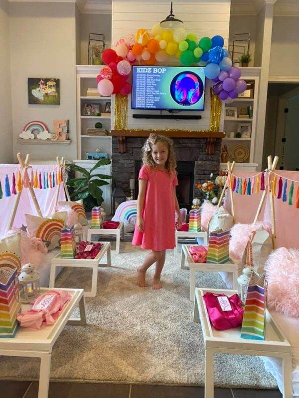 A young girl in a pink dress smiles at a boho rainbow theme birthday party set up with colorful decorations, a table with gift bags, and a tv screen displaying "kidz bop" in a cozy living room.