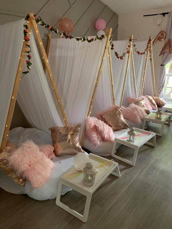 Indoor party setup with three cozy swing beds decorated with white drapes and floral garlands, complemented by pink cushions, fluffy throws, and small white tables with lanterns in a glam theme.