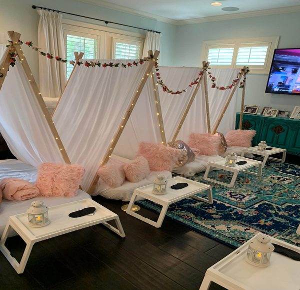 Cozy indoor glam theme setup with white Glamping Tent, pink fluffy cushions, fairy lights, and small lanterns on wood trays, in a living room with a decorative rug and TV in the background