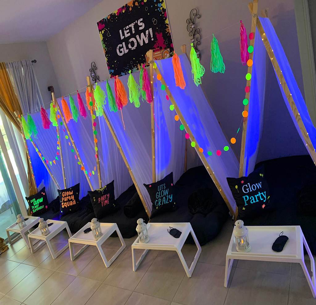 A festive room setup for a Glow Theme sleepover party featuring a banner that reads "let's glow," neon decorations, pillow forts with black cushions, and small white tables with glow sticks.