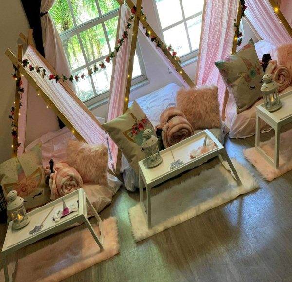 Cozy indoor glamping setup with two Glamping teepees adorned with lights, surrounded by fluffy pillows, pink blankets, and wooden tray tables with decorative items.