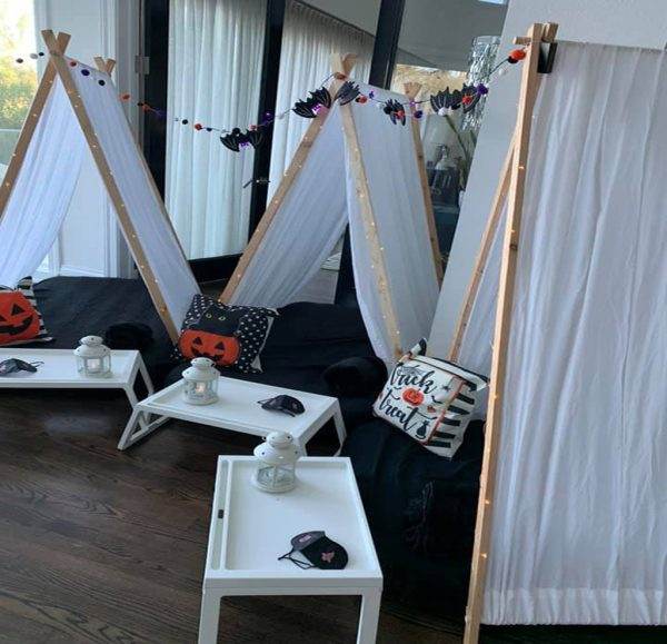 Three teepee tents decorated with Halloween-themed items including pumpkins and "trick or treat" signs, set up for a festive sleepover party, complete with small lanterns and themed cushions.
