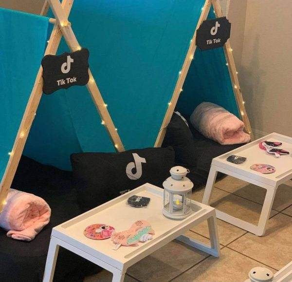 A cozy TikTok-themed slumber party nook with a teepee tent made of wooden poles and teal fabric adorned with the TikTok logo, facing two small white tables with lanterns, stickers, and