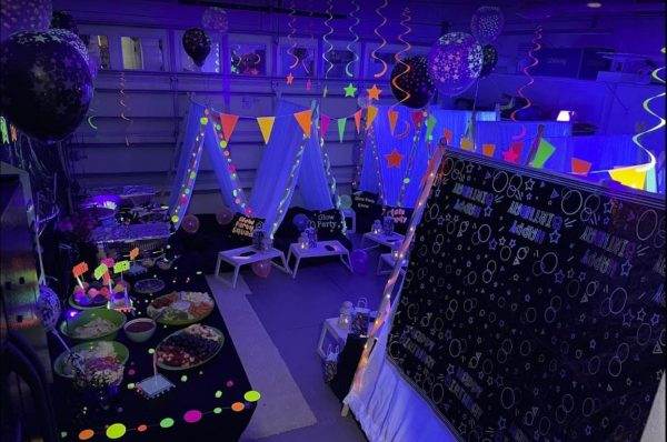 A vibrant Teepee Party setup featuring a blacklight theme with glowing decorations, balloons, and a chalkboard covered in drawings. festive tents, colorful banners, and a table full of food add to the fun atmosphere.