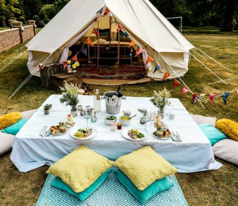 A luxurious bell tent glamping setup in a grassy field featuring an elaborately decorated tent with open flaps and a cozy picnic area in front, complete with cushions, a picnic table set with food, and champagne.