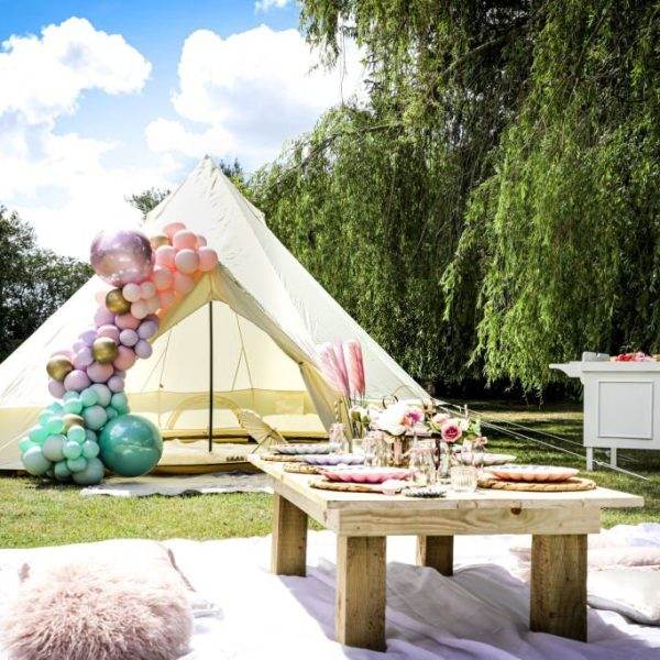 A luxurious outdoor setting featuring a Bell tent adorned with colorful balloons, surrounded by a lush green lawn under a clear blue sky. a picnic table and birthday cakes for children's party enhance the festive ambiance.
