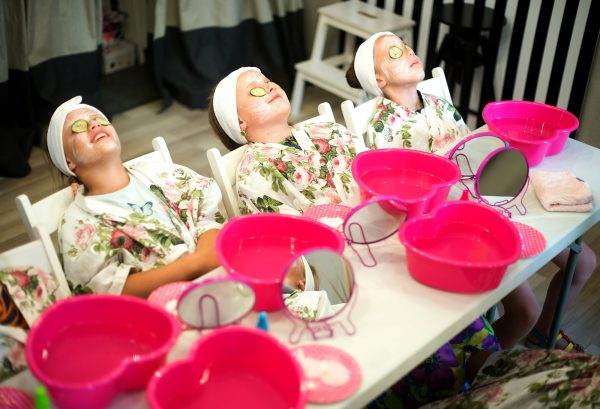 Three girls enjoying the excitement of a birthday party at a children's spa, wearing floral robes and headbands with face masks and cucumber slices over their eyes, relaxing with pink bowls and towels in front of them.