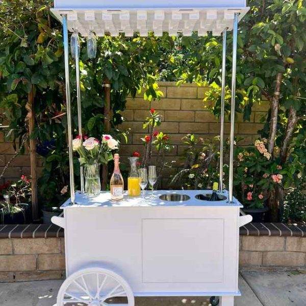A charming outdoor beverage cart adorned with flowers, a champagne bottle, and two glasses, standing in front of a lush green hedge and an XL white Outdoor Games Addon perfect for a party, complete with a bottle of wine on a sunny day.