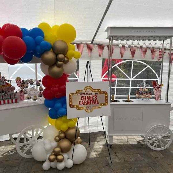A festive carnival-themed party setup under a white tent with a balloon cart adorned with patriotic red, white, and blue XL Outdoor Games Addon, a welcome sign reading "welcome to Chase's carnival," and an XL outdoor games station decorated with striped details.