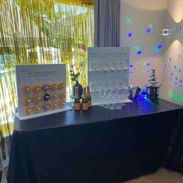 A party setup with a table displaying a donut wall labeled "donuts", an addon champagne bottle stand labeled "champagne", glasses, festive lights, and XL Outdoor Games Addon.