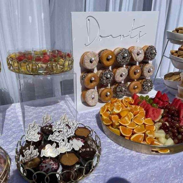 A dessert table with a variety of treats including a stack of doughnuts labeled "donuts", trays of sliced oranges, and plates of assorted pastries and XL Outdoor Games-themed fruit.