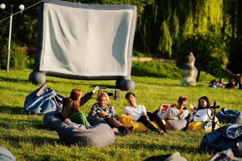 A group of young adults enjoys an Open air cinema party. they are seated on large bean bags, chatting and laughing, with a large blank projector screen in the background.