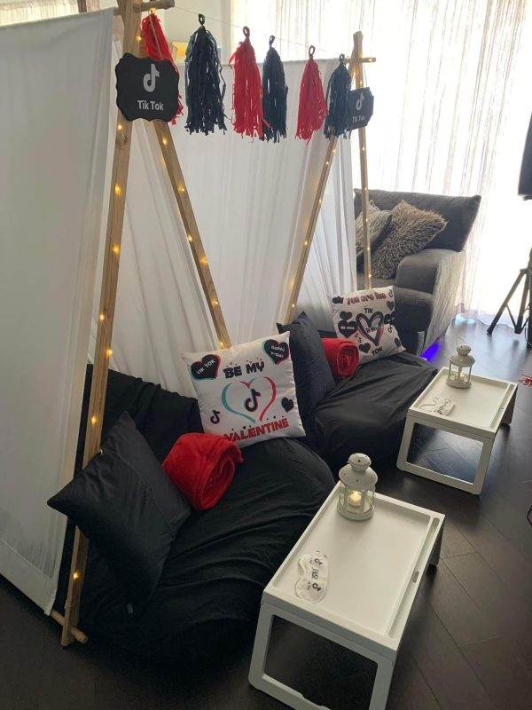 A cozy indoor relaxation nook with black cushions and decorative pillows, enhanced with string lights along a wooden frame, a white side table, and Tik-Tok Teepee themed party decorations.