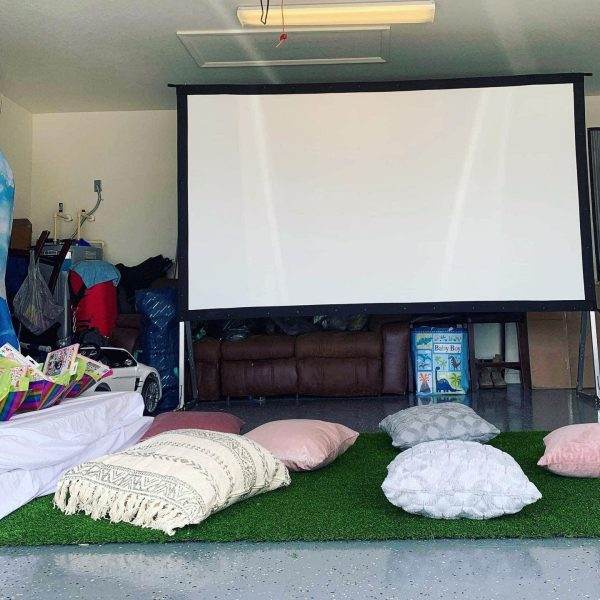 A glamping party setup in a garage with a large screen, teepee rentals, throw pillows on artificial grass, and various items stored around the space.