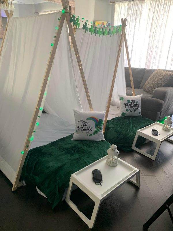 A cozy dino birthday party slumber party sleepover tepee rental decorated for St. Patrick's Day with a green theme, shamrock decorations, and a festive pillow. Small tables with a teapot and mugs are nearby.