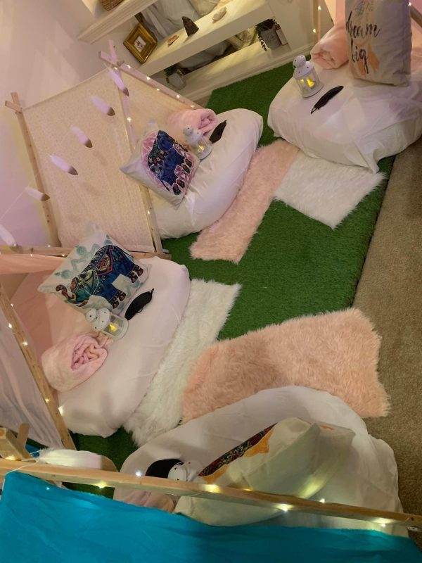 A cozy indoor Teepee Party w/ Fake Grass rug rental setup with two white bean bag chairs, fluffy pink and white pillows, a small tent frame with hanging fairy lights, and a green artificial grass carpet.