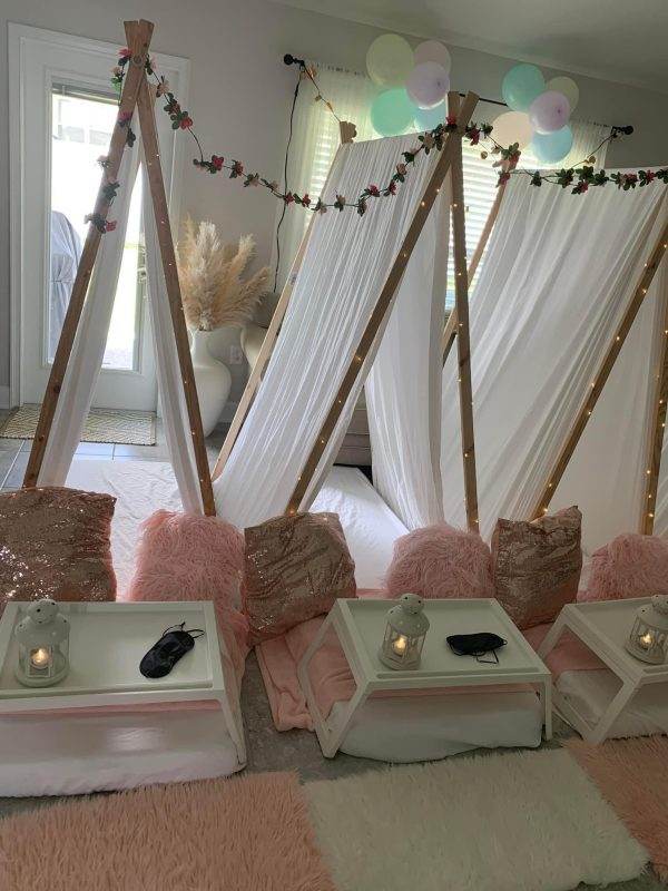 Pink and white teepee tents create a fun and whimsical party atmosphere in a living room featuring sleepover tent rentals, adorned with fairy lights and floral garlands, surrounded by pink and white cushions and small white tables with candles.