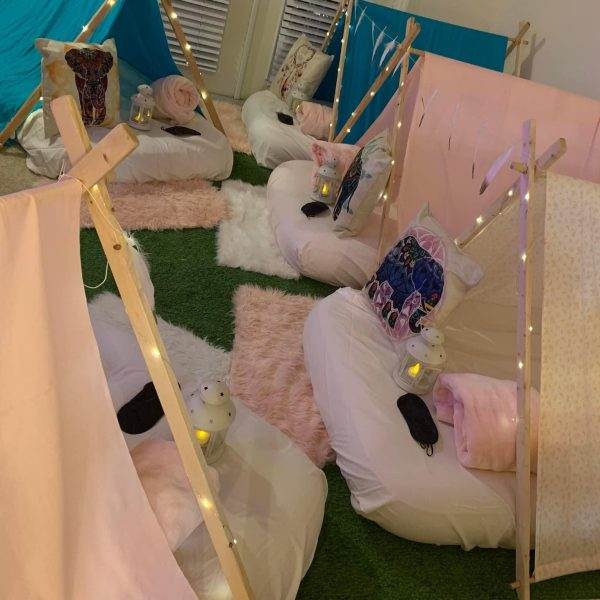 Indoor camping setup with small Sleepover Tent Rentals made of sticks and lights, surrounded by cushions and blankets on artificial grass.