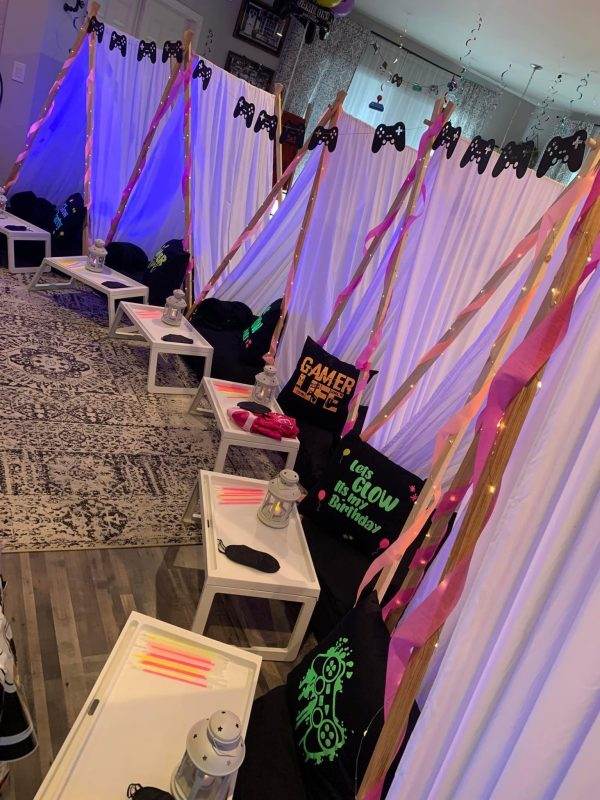 A Glow party-themed party room with rows of white gaming stations, each draped with pink and gold ribbons, featuring gamer signs and glow-in-the-dark elements on black cloths, complemented by sleepover