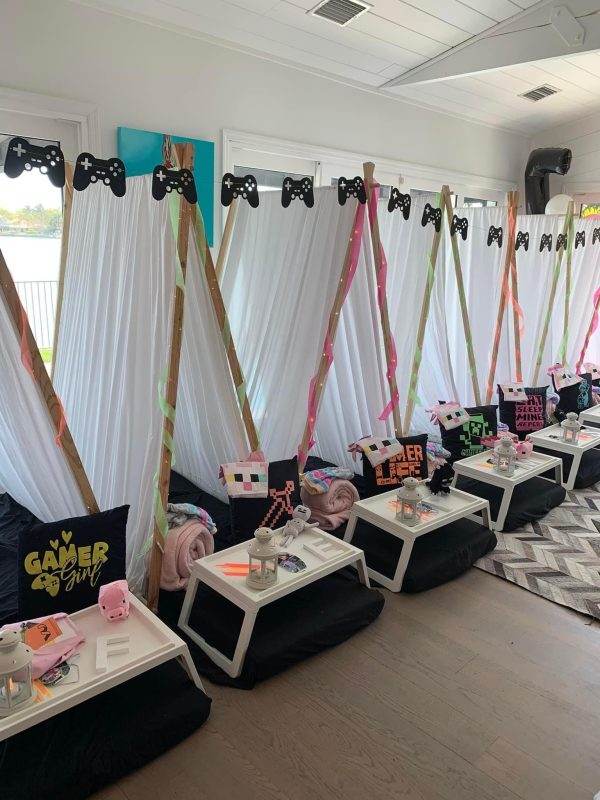 A party room with a gaming theme, decorated with video game controllers and ribbons hanging from the ceiling, and equipped with sleepover tent rentals and low tables for guests.
