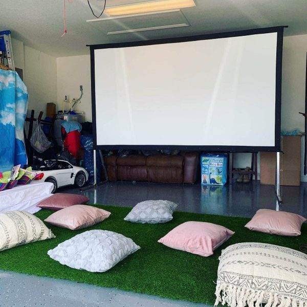 A garage converted into a cozy movie viewing area with a large screen, scattered cushions on artificial grass, a leather sofa, and various sleepover tents.