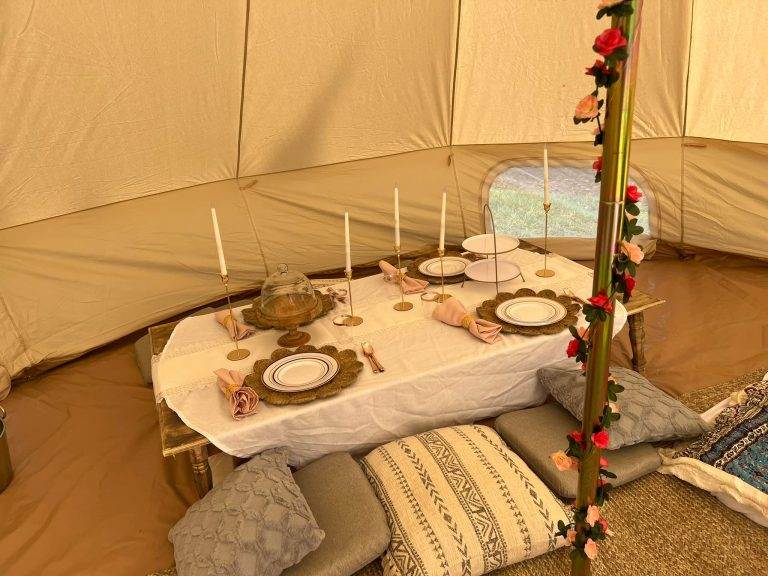 Elegant dining setup inside a spacious teepee tent, featuring a set table with plates, candles, and floral decorations, surrounded by plush pillows and rich textures.