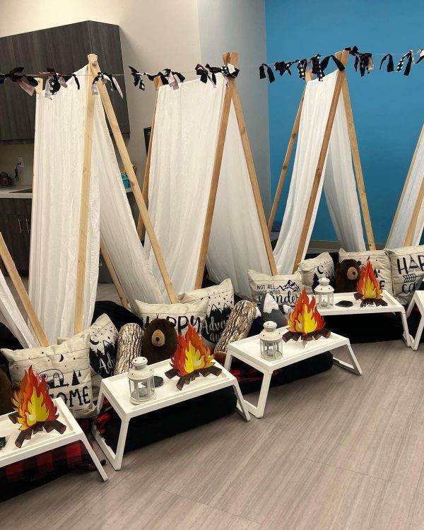 Sentence with product name: A teepee tent is set up in a room, transforming it into a cozy party space with four canvas tents, each featuring an artificial campfire, decorative pillows, and a small serving tray on a rug.