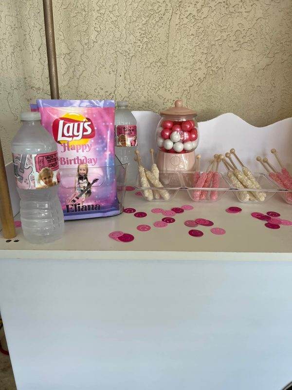 A birthday-themed display with custom "happy birthday" potato chips, pink candies in a jar, bubblegum on sticks, a water bottle, and scattered pink confetti against a Large Greenery Photo Backdrop with Neon Sign.