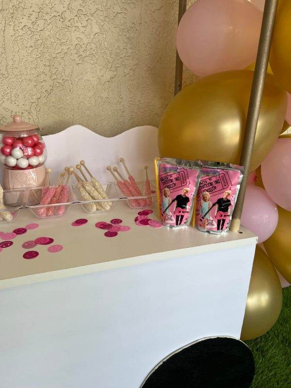 Party treats on a white table, including pink-wrapped lollipops and various candies, with a Large Greenery Photo Backdrop with Neon Sign and pink balloons in the background.