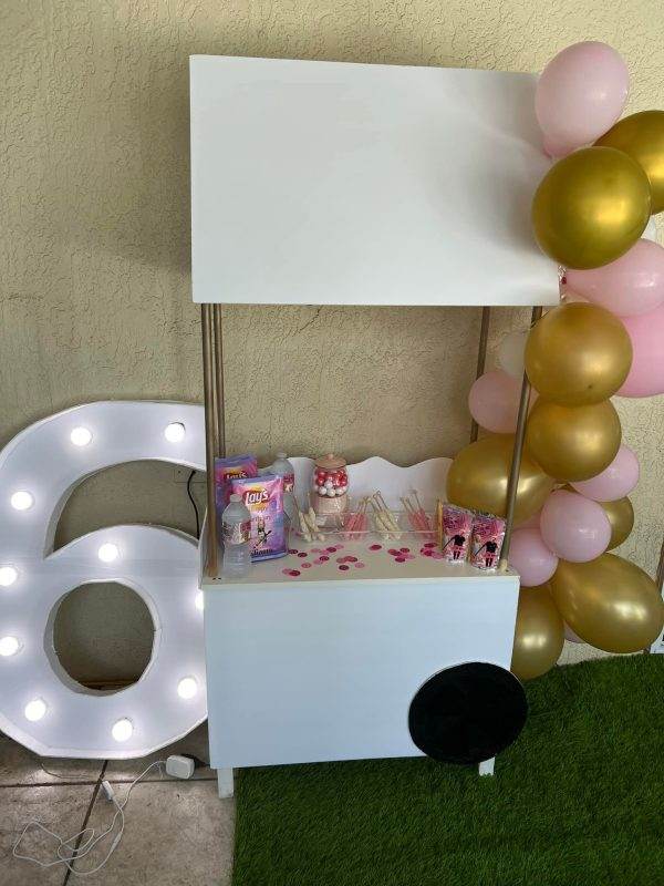 A kiosk-style booth decorated for a party with a large light-up product name, pink and gold balloons, and various party favors on a shelf under a white canopy.