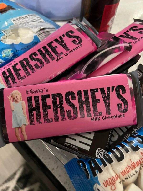 A personalized Hershey's milk chocolate bar with the name "Elliana" printed on the label, surrounded by other Hershey's bars in differing colors and designs, set against a Large Greenery Photo Backdrop with Neon Sign featuring Marilyn Monroe.