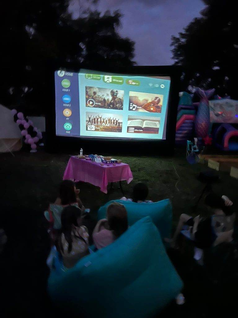 Children seated on 292547489_1435129800244540_6704579261717122892_n and chairs outdoors at dusk, watching a movie projected on a large screen, with a snack table in front.