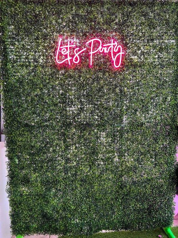 A Large Greenery Photo Backdrop with Neon Sign saying "let's party" in pink cursive script.
