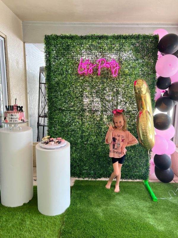 A little girl standing in front of a Teepee Party setup featuring a Large Greenery Photo Backdrop with Neon Sign, surrounded by pink and black balloons and treat-filled pedestals.