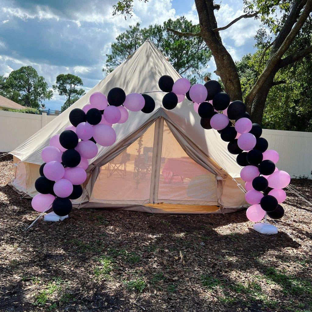 A large 293377074_1435127180244802_8023254540460888526_n tent set up outdoors under a sunny sky, adorned with an arch of black and pink balloons at the entrance.