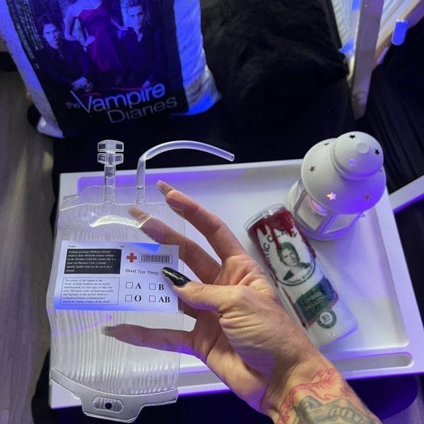 A person with a tattooed arm holding an IV bag with Juice Blood Bags Birthday Party Add-on marked on it, with a "the vampire diaries" bag and other miscellaneous items in the background.