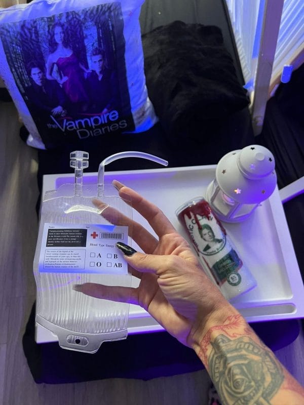 A person's tattooed hand holding a "Juice Blood Bags Birthday Party Add-on" over a table with a "The Vampire Diaries" bag, a wallet, and a small projector.