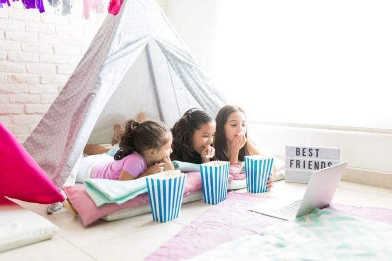 Three young girls lie on pillows, enjoying a slumber party, watching a laptop with popcorn beside them under a tent made of blankets.
